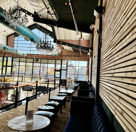 Loft-style second-floor lounge with industrial-chic vibe: crystal chandeliers, teal exposed ductwork, wood-slat accent wall, velvet sofas, round marble tables and floor-to-ceiling windows overlooking the street.