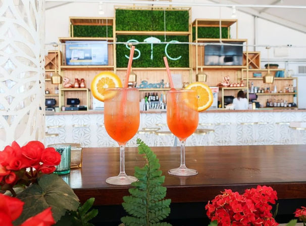 Two sunny orange cocktails garnished with orange slices and straws on a wooden bar counter in a bright beachside bar interior with tropical plants and red flowers in the foreground.