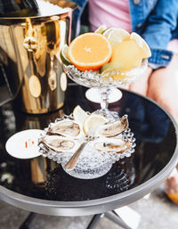Crystal bowls on a black bistro patio table holding fresh oysters on ice with lemon wedges and a second bowl of grapefruit, lime and lemon slices next to a gold champagne bucket