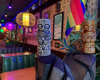 Two carved tiki mugs atop a wooden carving in a colorful tiki-bar interior with rattan pendant lights, green vinyl booths, framed wall art and a rainbow pride flag under purple and multicolored neon lighting.