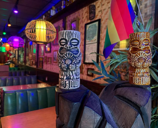 Two carved tiki mugs atop a wooden carving in a colorful tiki-bar interior with rattan pendant lights, green vinyl booths, framed wall art and a rainbow pride flag under purple and multicolored neon lighting.