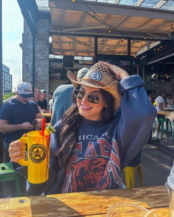 Smiling woman in sunglasses and a straw cowboy hat holding a large yellow mug with a garnished cocktail on a sunny urban patio bar with string lights and a lively crowd.
