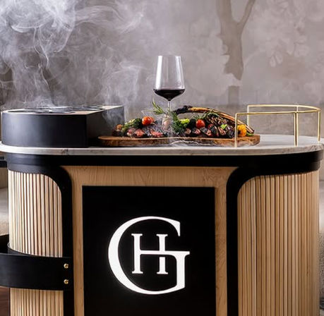 Marble-topped tableside cart with steaming grilled meats and roasted vegetables on a wooden board, glass of red wine, and illuminated emblem — upscale restaurant presentation.