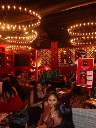 Cozy upscale cocktail lounge interior at night with warm red lighting, large circular chandeliers, patterned wallpaper, plush sofas and groups of people socializing