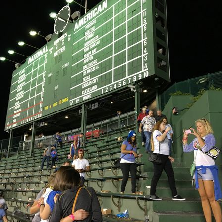 Nighttime green manual baseball scoreboard and clock towering over tiered stadium bleachers with fans exiting, taking photos, and walking down steps in a lively outdoor ballpark scene.