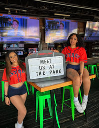 Two women in red Astros crop jerseys pose with a tabletop sign reading "MEET US AT THE PARK" inside a lively outdoor sports bar with bright green stools and multiple TVs.