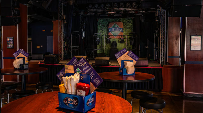 Empty live-music bar interior with round wooden tables and stools, dark concert stage with lighting in the background, and Bud Light condiment caddies holding napkins and ketchup on foreground tables, ready for a show.