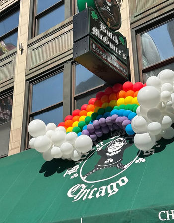 Colorful rainbow balloon arch with white cloud balloons decorating a green downtown Chicago storefront awning beneath a vintage pub sign, festive Pride display.