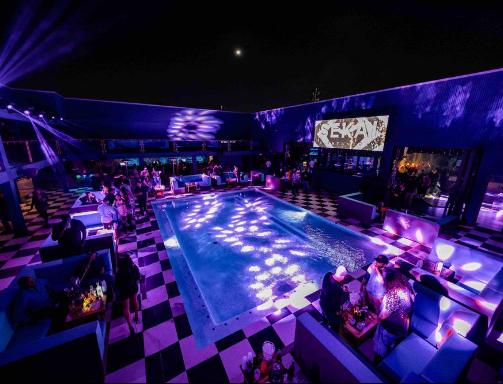 Vibrant rooftop nightclub at night featuring a glowing rectangular pool, purple and blue lights reflecting on the water, black-and-white checkered floor, booth seating and groups of people socializing around tables.