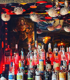 Eclectic cocktail bar interior: crowded shelf of assorted liquor bottles under warm red lights, ceiling adorned with hanging vinyl records and spiky dried pufferfish ornaments, with a dramatic armored portrait framed in the background.