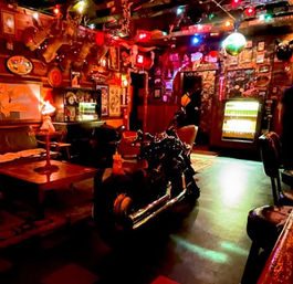 Dimly lit retro bar interior with a chrome motorcycle parked inside, eclectic wall art and trophies, string lights, disco ball, vintage vending machine and cozy seating.