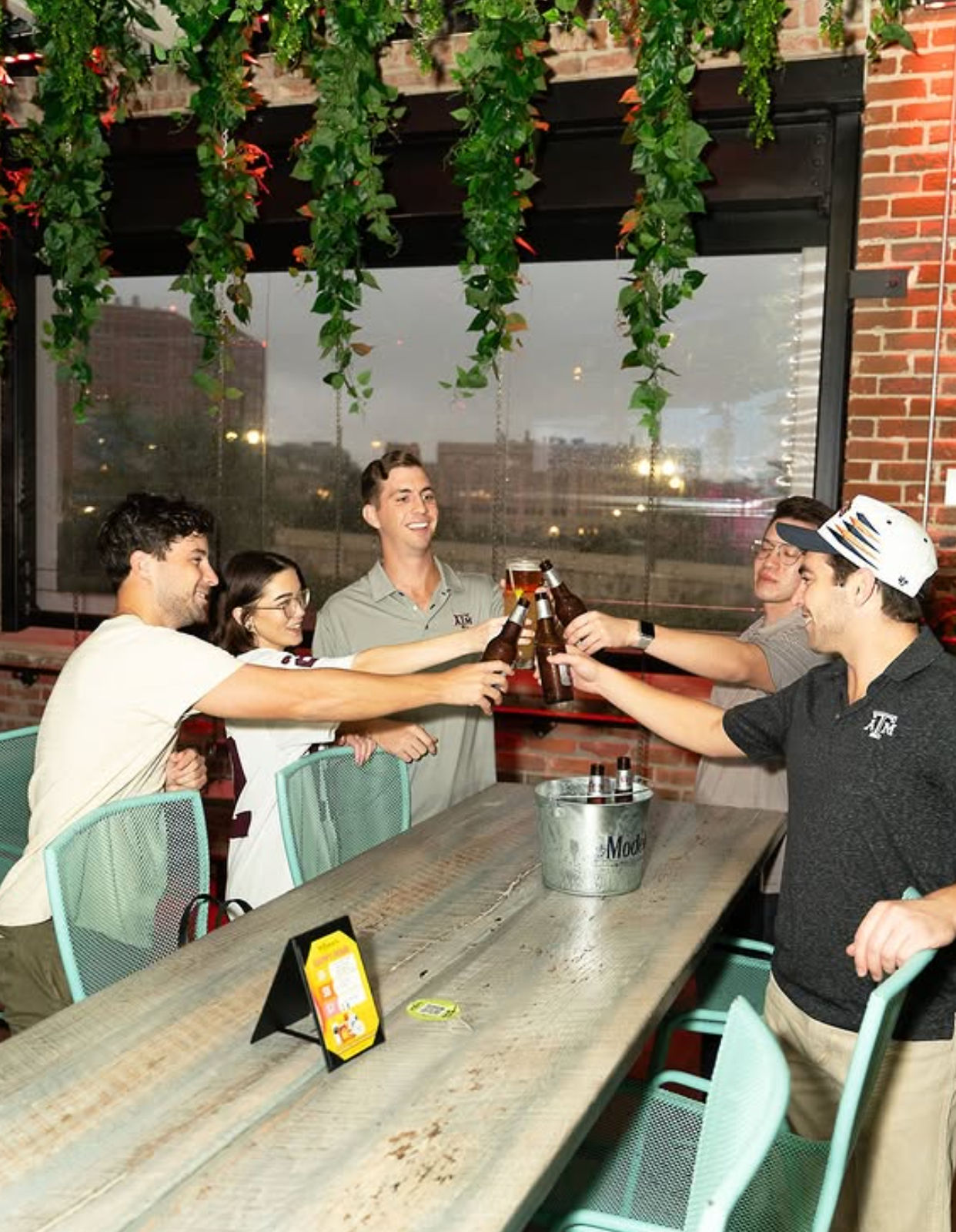 Five friends clinking beer bottles around a long wooden table on an urban rooftop patio with hanging plants and exposed brick, cheering and smiling.