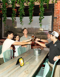 Five friends clinking beer bottles around a long wooden table on an urban rooftop patio with hanging plants and exposed brick, cheering and smiling.