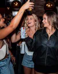 Three friends laughing and toasting with drinks in a dimly lit bar, one holding a slim canned beer while others raise cocktails against a warm pendant-light and stone wall backdrop.
