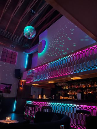 Neon-lit cocktail lounge interior with a hanging disco ball casting teal and purple speckles, geometric honeycomb balcony and backlit bar with stools and bottles