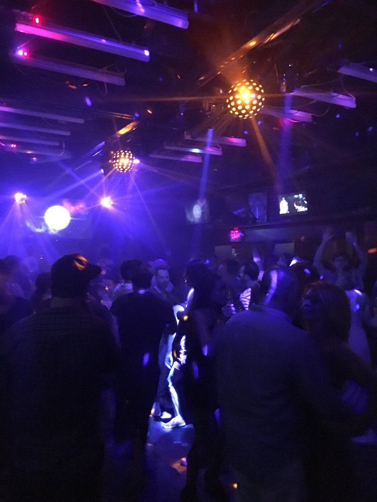 Crowded indoor nightclub dance floor with purple and blue disco lights and glowing mirror balls, silhouettes of people dancing under ceiling spotlights.