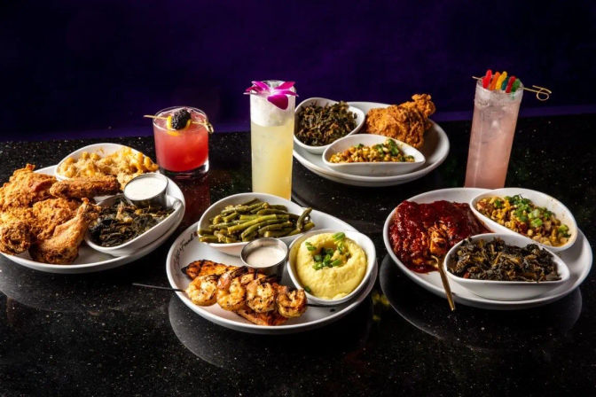 Vibrant spread of Southern comfort food on a black table: crispy fried chicken, BBQ ribs, shrimp skewers, mashed potatoes, green beans, collard greens, mac and cheese and colorful cocktails.