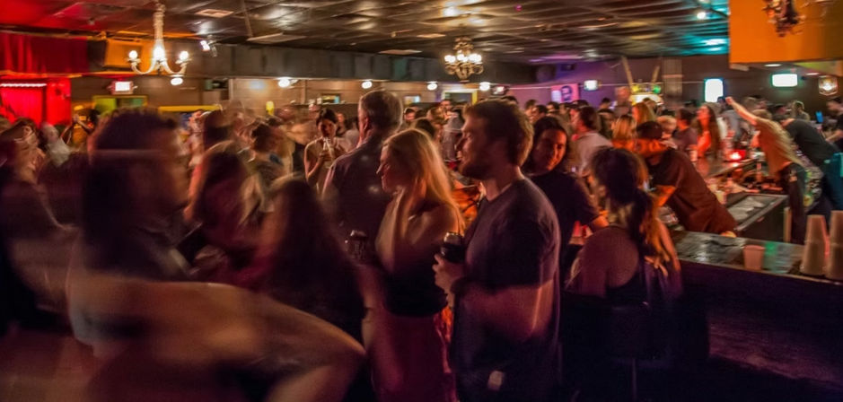 Packed dimly lit bar at night with warm chandeliers, buzzing crowd, blurred dancers and patrons mingling around a busy bar — lively nightlife scene.