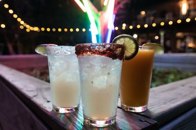 Three cocktails on a wooden outdoor patio at night: two pale ice-filled drinks and an orange cocktail, center glass rimmed with chili powder and garnished with a lime wheel, colorful neon lights and bokeh string lights in the background.