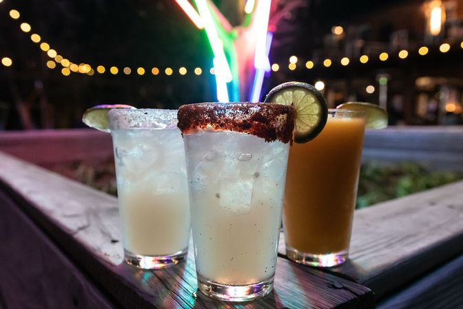 Three cocktails on a wooden outdoor patio at night: two pale ice-filled drinks and an orange cocktail, center glass rimmed with chili powder and garnished with a lime wheel, colorful neon lights and bokeh string lights in the background.