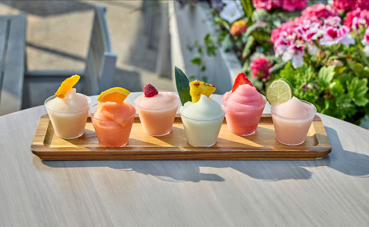 Six colorful frozen cocktails on a wooden tasting board on an outdoor patio table, each garnished with citrus peel, orange slice, raspberry, pineapple leaf, strawberry, and lime wheel