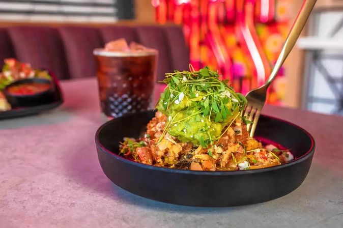 Brunch hash bowl topped with smashed avocado and microgreens, fork digging in on a restaurant table with an iced cocktail and colorful interior in the background.