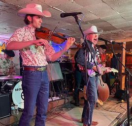 Toe-tapping country band performing in a small indoor venue: fiddler in a cowboy hat, singer-guitarist with white hat, upright bass, drummer and colorful stage lights.