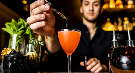 Bartender adding bitters with a dropper to a vibrant orange craft cocktail in a coupe glass on a dimly lit bar, surrounded by jars of mint, fruit garnishes, and bottles.