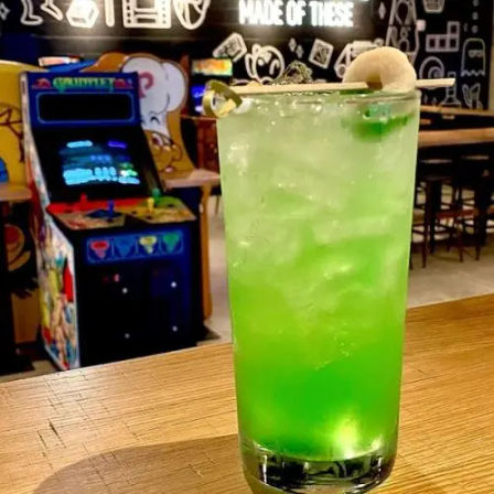 Neon green highball cocktail with crushed ice, lime slice and lychee skewer on a wooden bar top, blurred retro arcade machine and bar seating in the background