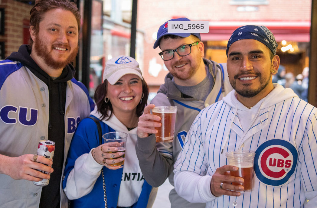 Four smiling friends in blue-and-white baseball jerseys and caps holding plastic cups of beer at an outdoor bar or tailgate