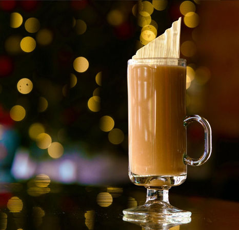 Festive holiday latte in a tall glass mug with a wafer garnish, warm golden Christmas-light bokeh and reflective surface
