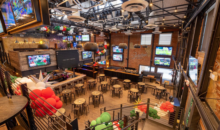 Spacious industrial loft-style sports bar interior with exposed brick walls, high metal ceilings, mezzanine seating, multiple large TV screens showing sports, wooden round tables and stools, central bar area and red, white and green balloon decorations — festive, game-day ready.