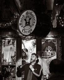 Black-and-white photo of a male stand-up comedian holding a microphone on stage inside a cozy Irish-style pub in Chicago, rustic wood interior, string lights, and logo-adorned merchandise displayed behind him.
