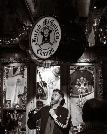Black-and-white photo of a male stand-up comedian holding a microphone on stage inside a cozy Irish-style pub in Chicago, rustic wood interior, string lights, and logo-adorned merchandise displayed behind him.