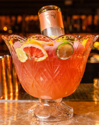 Crystal punch bowl of pink citrus cocktail with grapefruit, lime and lemon slices and an inverted bottle on a bar counter