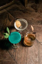 Three craft cocktails on a rustic wooden bar table — bright turquoise coupe with mint, clear martini with citrus peel and olive, and amber old-fashioned with a large ice cube.