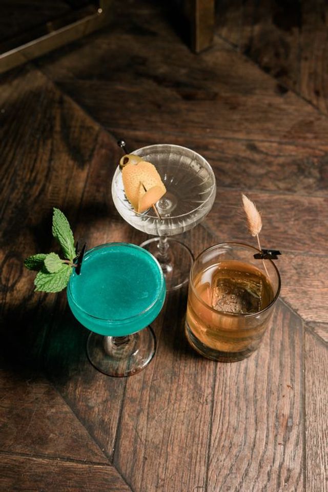 Three craft cocktails on a rustic wooden bar table — bright turquoise coupe with mint, clear martini with citrus peel and olive, and amber old-fashioned with a large ice cube.