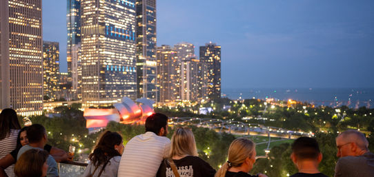 Rooftop crowd overlooking Chicago skyline at dusk, glowing skyscrapers, the illuminated metallic band shell in a waterfront park and Lake Michigan in the background.