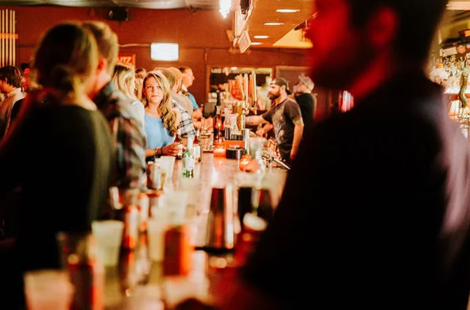 Busy neighborhood bar interior at night with patrons gathered along a long illuminated bar and a bartender pouring drinks under warm orange lighting