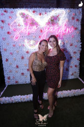 Two smiling women posing at a photo-op in front of a pink neon butterfly script sign on a white-and-pink rose wall backdrop at a lively event.