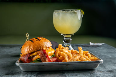 Bacon cheeseburger with melted cheddar and tomato on a toasted brioche bun, crispy seasoned fries on a metal tray, and a salted margarita in a goblet — American casual dining plate.