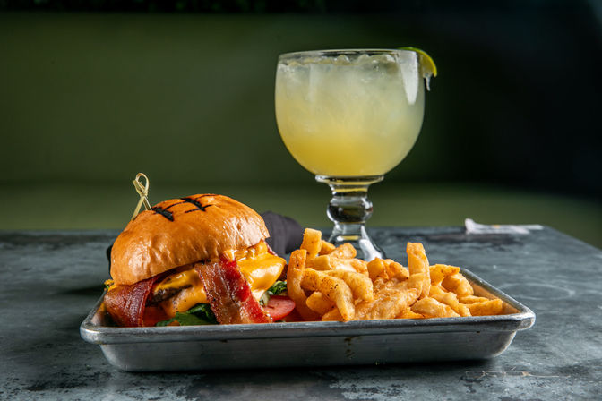 Bacon cheeseburger with melted cheddar and tomato on a toasted brioche bun, crispy seasoned fries on a metal tray, and a salted margarita in a goblet — American casual dining plate.