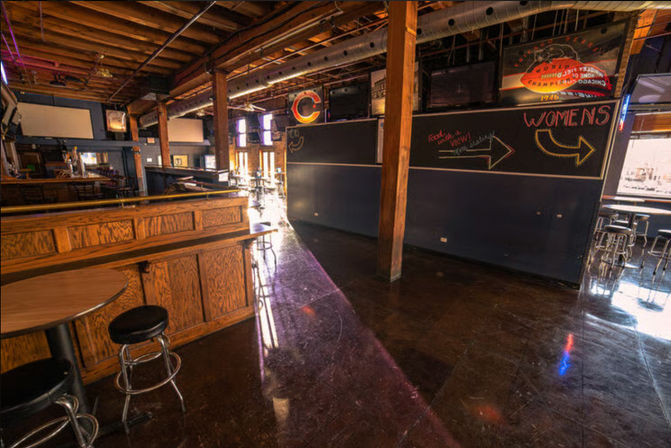 Sunlit industrial-style bar interior with a long wooden bar, round tables and metal stools, exposed timber beams, polished dark floor, and chalkboard arrows pointing to the women's restroom.