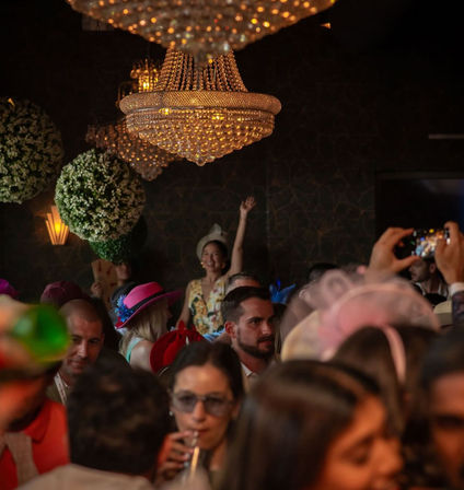 Festive indoor crowd in an elegant venue beneath sparkling crystal chandeliers, guests wearing colorful hats and fascinators, one person waving while others sip drinks and take photos.