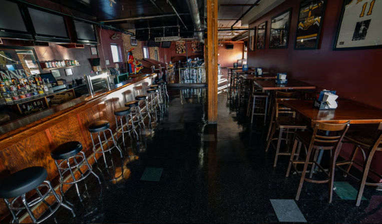 Empty sports-style bar interior with long wooden counter and row of metal swivel stools, high-top tables and chairs along maroon walls adorned with framed jerseys and photos, glossy black floor reflecting light.