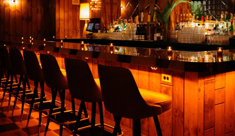 Cozy wood-paneled cocktail bar with glossy black countertop, row of leather bar stools, warm amber under-counter lighting and shelves of bottles creating an intimate nighttime vibe