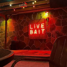 Red neon sign reading "LIVE BAIT" mounted on a stone wall of a dim outdoor patio with string lights, a chair and table, and warm red ambient lighting.