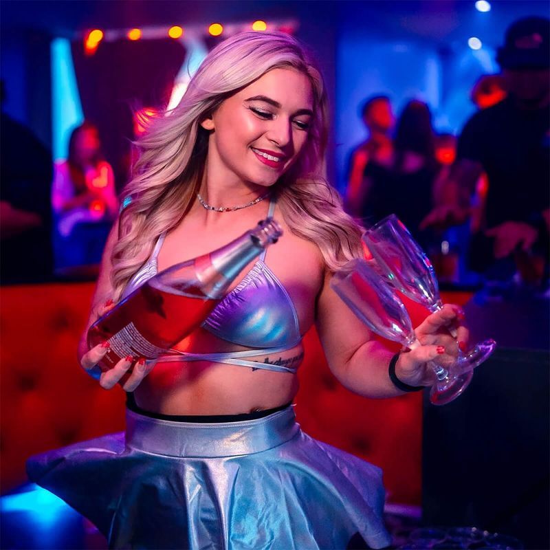 Smiling woman in metallic lilac outfit pouring sparkling rosé into two wine glasses in a colorful nightclub setting