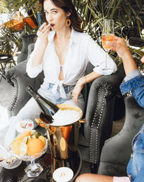 Stylish woman in a white tied shirt nibbling a bite while a friend toasts rosé; champagne bottle in a gold ice bucket, citrus bowl and velvet studded armchairs on a leafy outdoor patio brunch.