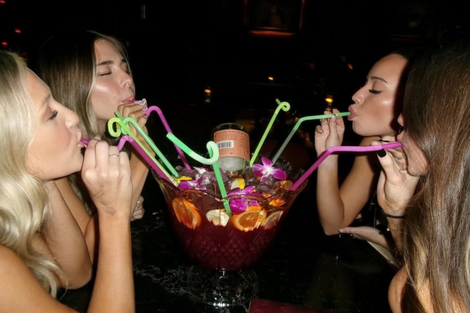 Four women sipping from a giant tropical punch bowl through colorful bendy straws, garnished with citrus slices and orchids in a dimly lit bar setting.
